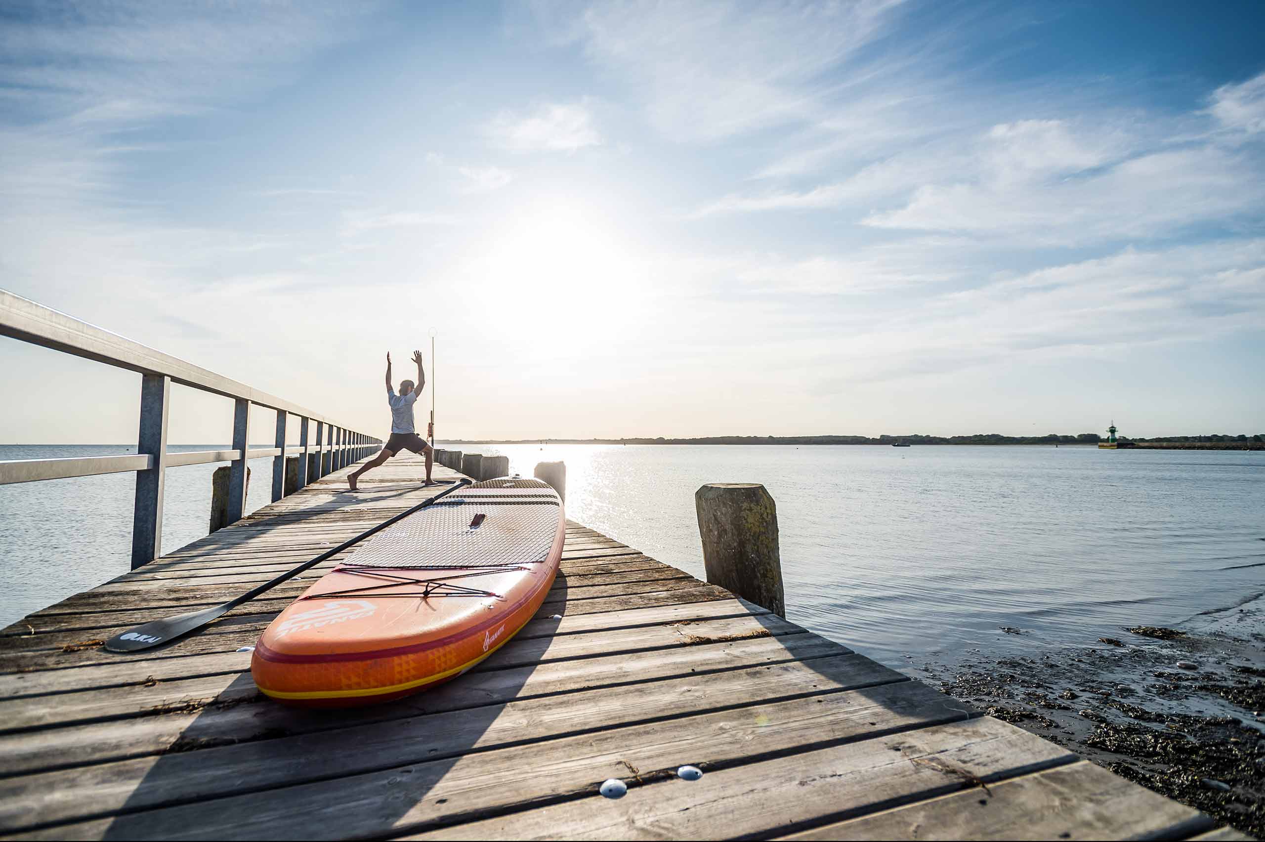 Mann macht Yoga auf einem Steg, im Vordergrund das SUP-Board